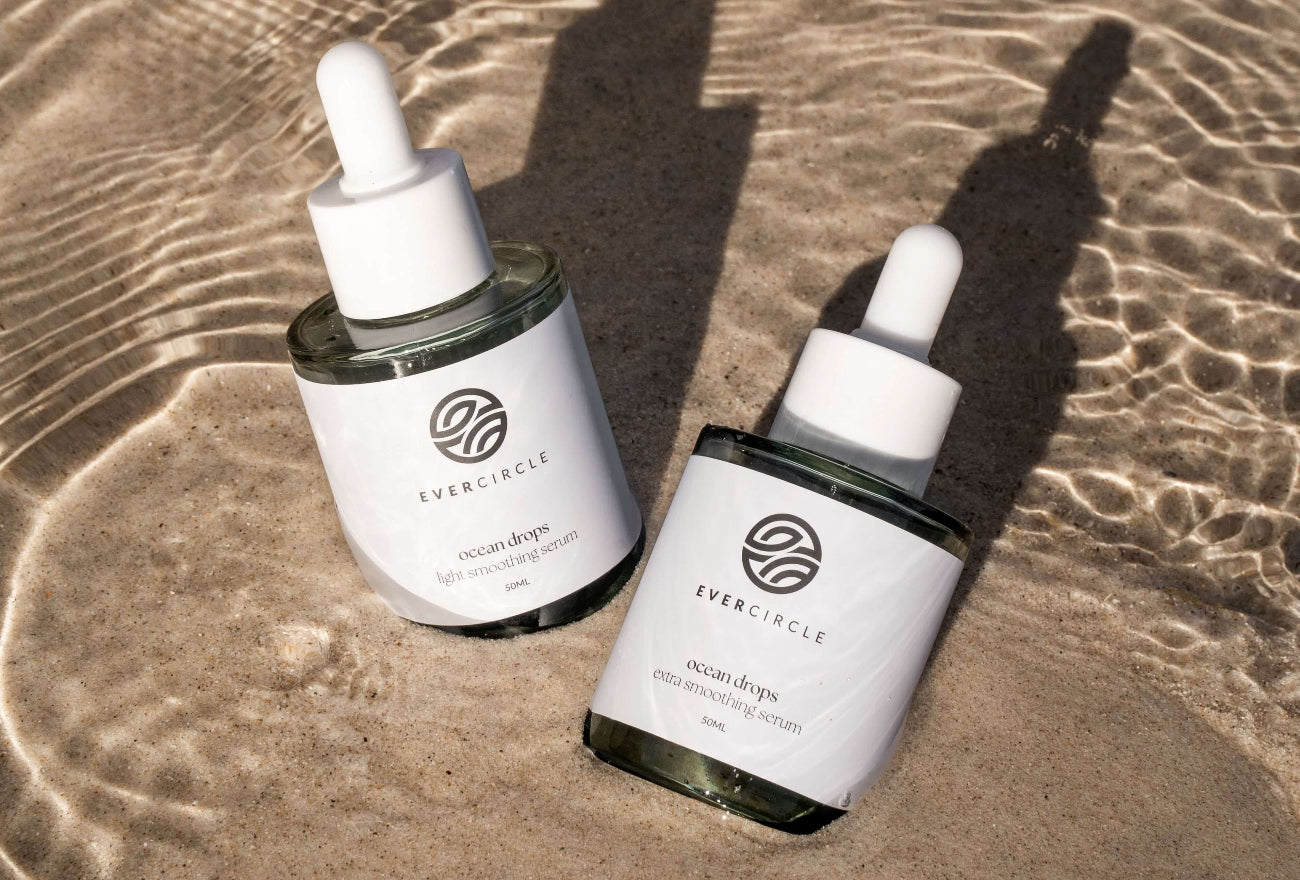 Two Beach hair protection oil bottles by Evercircle sitting on the sand with saltwater rippling around them.