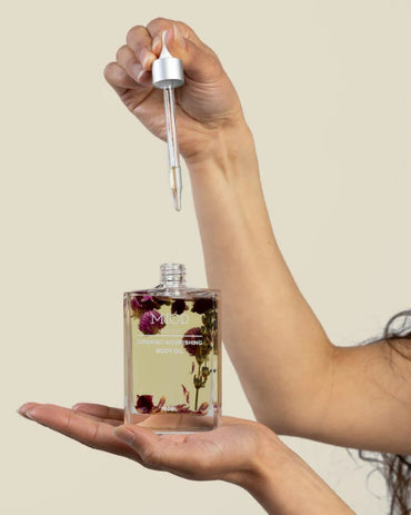 Hand holding a dropper over a bottle of natural body oil with a beige background