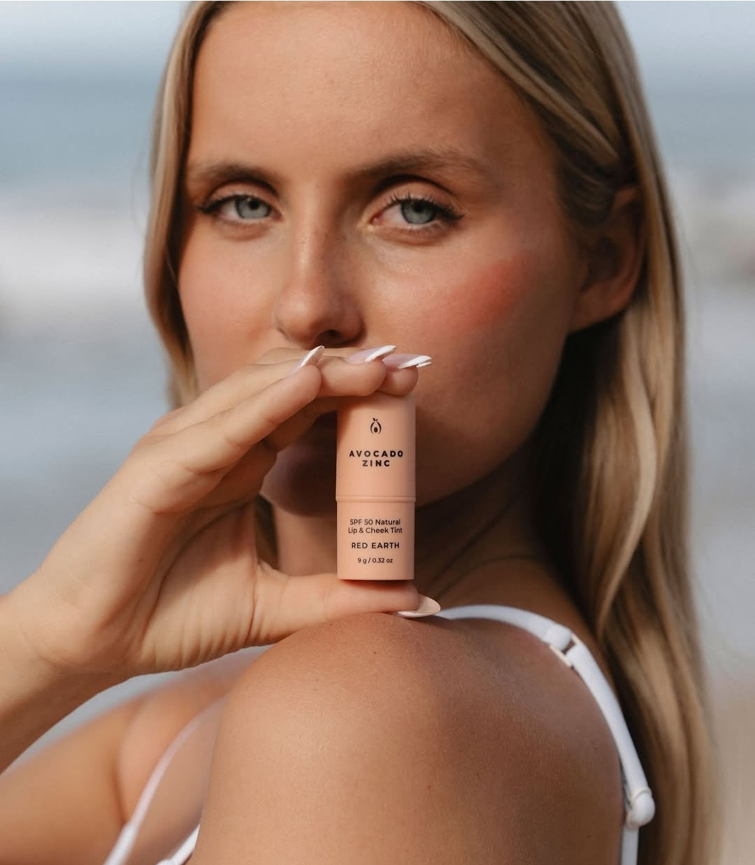 Women holding a tube of avocado zinc lip and cheek tint SPF make-up to her face. 