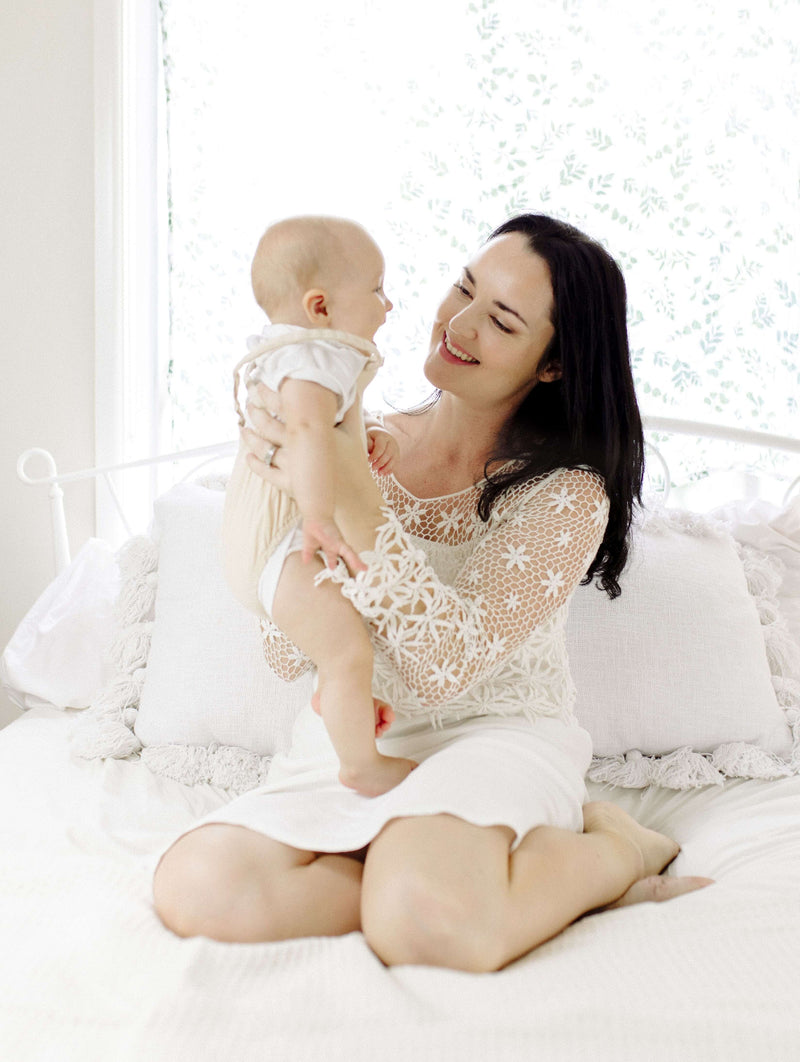 Woman holding a baby in a white lace dress on a bed with white bedding.
